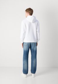 Sweatshirt blanc avec capuche, associé à un jean bleu clair, présentant une coupe décontractée et un style à cinq poches, sur fond uni.