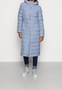 Abrigo acolchado azul claro con cierre de cremallera, mangas largas, capucha y patrón acolchado vertical, llevado sobre vaqueros oscuros y zapatillas grises.