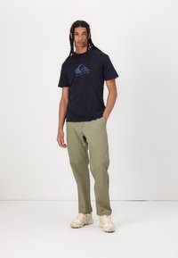 Marinebomulls t-skjorte med rund hals og Quiksilver-logo, kombinert med lysegrønne bukser og kremfargede sneakers, vist på en nøytral bakgrunn.