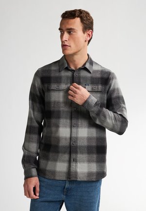 Camisa de franela a cuadros en gris y negro con diseño de botones, mangas largas y dos bolsillos en el pecho. Textura suave y ajuste casual.