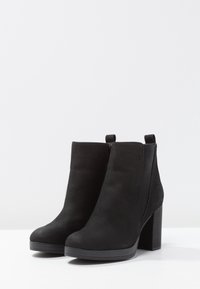 Bottes noires en daim à la cheville avec un haut talon carré, des panneaux élastiques sur les côtés et des languettes pour un enfilage facile. Texture lisse, design minimaliste.
