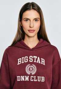 Sudadera color burdeos con el texto "BIG STAR DNM CLUB" en blanco, con un detalle de logo y una tela suave y texturizada. Diseño con capucha, estilo informal.