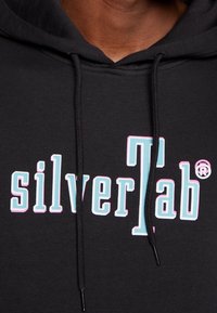 Svart hoodie med en djärv "silverTab"-logotyp i ljusblå och rosa detaljer, med dragsko och en strukturerad tyg.