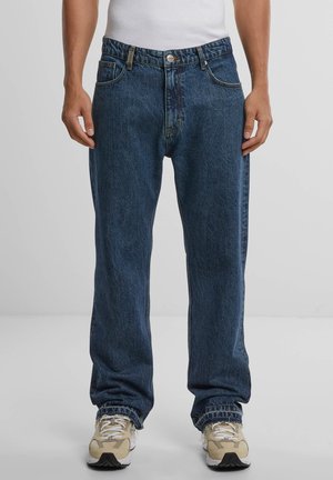 Jean en denim bleu avec une coupe ample, cinq poches, une braguette à boutons et des coutures visibles. Portés avec des baskets beiges.
