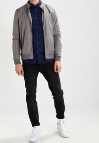 Lehká šedá bomber bunda s vysokým límcem, nošená přes tmavě modrou košili na knoflíky, spárovaná s černými džíny a bílými teniskami.