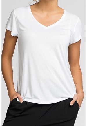 Kobieta ubrana w gładką, białą koszulkę z krótkim rękawem i dekoltem w serek, z rękami w kieszeniach, w połączeniu z czarnymi spodniami na neutralnym tle.