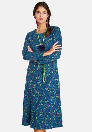 Ein langärmliges Kleid aus einem blauen Stoff mit einem lebhaften Blumenmuster in Grün und Lila, kombiniert mit grünen Perlen und dunklen Blumenaccessoires.