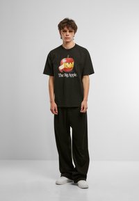 Camiseta de algodón negra con un gráfico colorido de una manzana y el texto "La Gran Manzana". Lleva pantalones negros holgados y zapatillas blancas.