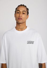 T-shirt en coton blanc à manches courtes, avec "LEGENDS" imprimé en noir sur le côté gauche. Encolure ras du cou, coupe décontractée.