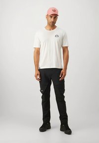 Balts īss piedurkņu T-krekls ar uzšuvumu, melnas kravas bikses ar kabatām un rozā cepure. Melni apavi ar teksturētu dizainu.