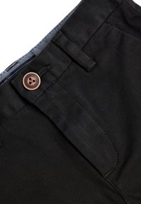 Schwarze Baumwollhosen mit einem Knopfverschluss vorne, Gürtelschlaufen und dezenter Textur, einschließlich einer Nahaufnahme der Nähdetails.