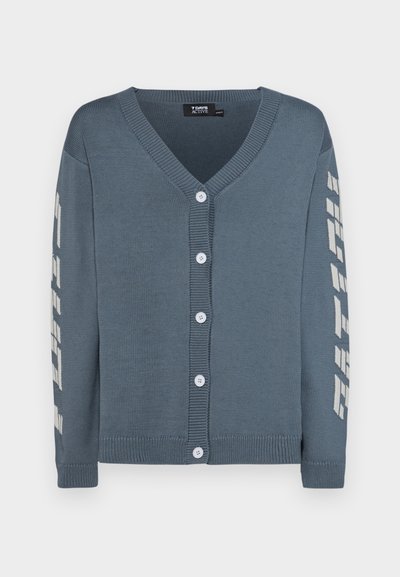 Cardigan boutonné en tricot doux gris-bleu, avec un col en V, cinq boutons blancs et un texte blanc contrastant le long de la manche gauche.