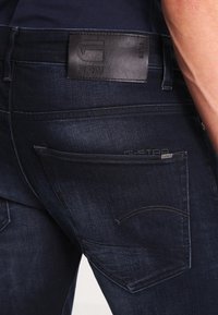 Mörkblå jeans i denim med en svart läderpatch från G-Star RAW och en synlig bakficka, bärs av en person som har en synlig hand och arm.