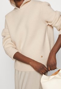 Helles beige gestricktes Sweatshirt mit Kapuze, gerippten Bündchen und Saum, mit einem kleinen goldenen Akzent auf der unteren linken Seite. Weiche Textur.