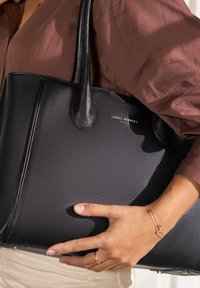 Schwarze Tasche aus genarbtem Leder mit doppelt tragenden Griffen und goldfarbener Logodekoration. Nahaufnahme, die die strukturierte Oberfläche und eine Hand zeigt, die die Tasche greift.