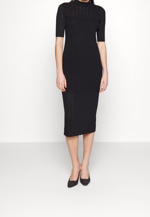 Kobieta w dopasowanej, czarnej, prążkowanej sukience midi z rękawami do łokci i czarnych szpilkach z ostrym noskiem, stojąca na białej podłodze.