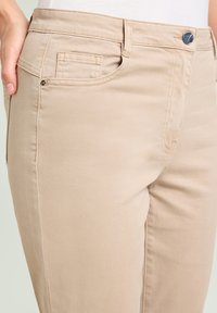 Pantalones de algodón beige con un corte ajustado, que cuentan con bolsillos frontales, un bolsillo para monedas y un cierre de botón metálico. Textura suave y detalles de costura.