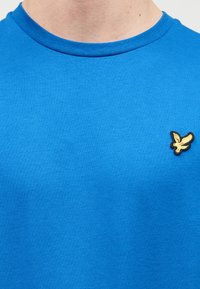 Μπλε βαμβακερό μπλουζάκι με στρογγυλή λαιμόκοψη και κοντά μανίκια. Κεντημένο λογότυπο πουλιού σε χρυσό χρώμα στην αριστερή πλευρά του στήθους. Μαλακή υφή.