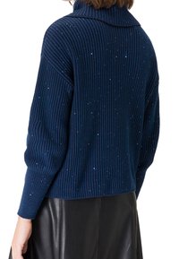 Mujer vistiendo un suéter de canalé azul oscuro con pequeños brillantes y una falda de cuero negra, vista de espaldas y desde la cintura hacia arriba.