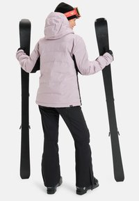 Padded light purple ski jacket with a black side, black ski pants, and ski equipment, viewed from behind against a white background.