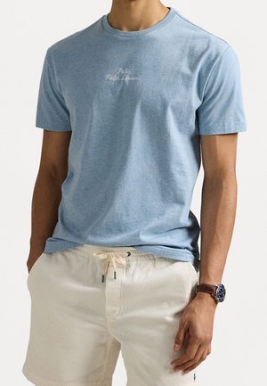Mężczyzna w jasnoniebieskiej koszulce Polo Ralph Lauren i kremowych szortach ze ściągaczem, z brązowym zegarkiem na lewym nadgarstku.