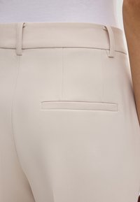 Pantaloni beige con una texture liscia, caratterizzati da un taglio dritto, una tasca posteriore e passanti per la cintura. Vista ravvicinata dell'area vita.
