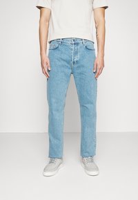 Man som bär ljusblå rakbenade jeans, vita sneakers och en kortärmad ljusbeige skjorta, stående mot en enfärgad vit bakgrund.