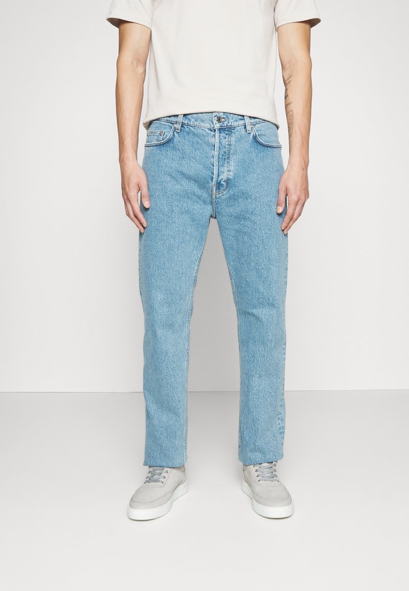 Man som bär ljusblå rakbenade jeans, vita sneakers och en kortärmad ljusbeige skjorta, stående mot en enfärgad vit bakgrund.