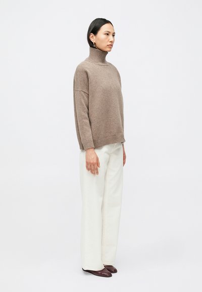 Γυναίκα που φοράει ζιβάγκο πουλόβερ σε χρώμα ταυπ, άσπρο παντελόνι με φαρδιά μπότα και σκούρα καφέ μυτερά παπούτσια, στέκεται μπροστά σε απλό φωτεινό φόντο.