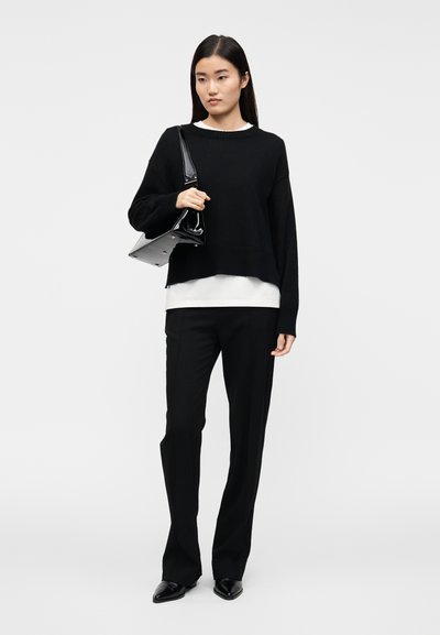 Pull noir oversize avec ourlet côtelé, porté sur un haut blanc. Le mannequin l'associe à un pantalon noir ajusté et un sac à main brillant.