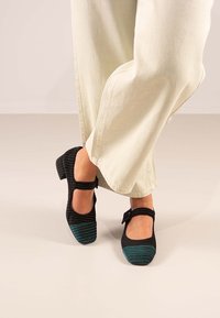 Zapatos de tela negros y verde azulado con una textura acanalada, que cuentan con una correa alrededor del tobillo y un tacón cuadrado, combinados con pantalones de color claro.