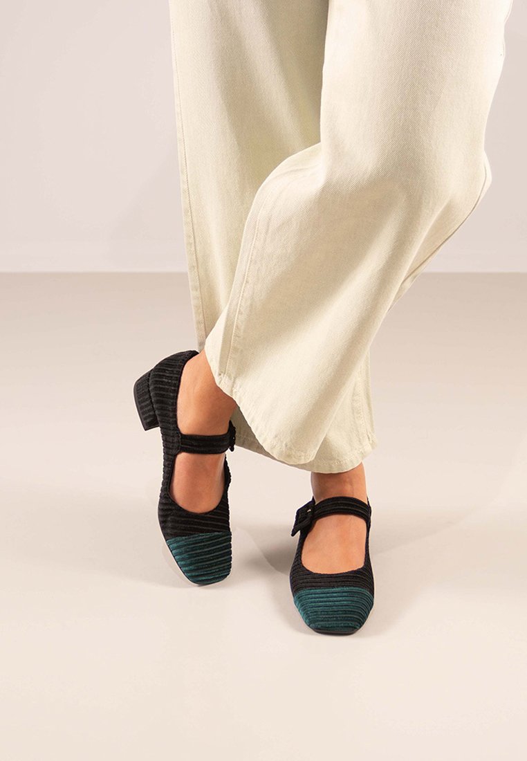 Zapatos de tela negros y verde azulado con una textura acanalada, que cuentan con una correa alrededor del tobillo y un tacón cuadrado, combinados con pantalones de color claro.