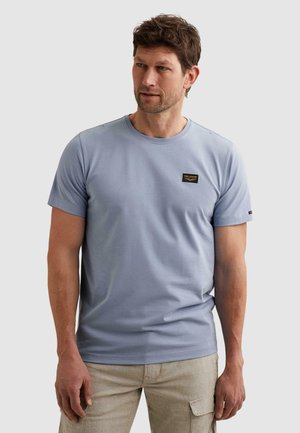 Muškarac u svijetloplavoj kratkih rukava majici s PME Legend oznakom na prsima i bež cargo hlačama, stoji na jednobojnoj pozadini.