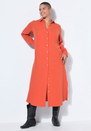 Hymyilevä lyhyttukkainen henkilö, joka pukeutunut oranssiin pitkähihaiseen nappimekkoon ja mustiin cowboy-saappaisiin, seisoo valkoista taustaa vasten.