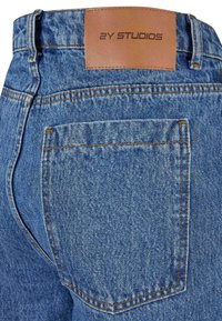 Poche arrière d’un jean en denim bleu avec une étiquette en cuir marron cousue au-dessus, embossée du texte « 2Y STUDIOS ».