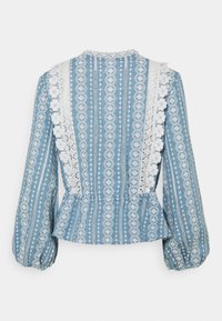 Blouse bleue à manches longues avec motifs géométriques verticaux blancs, bordure en dentelle blanche et détails volantés à la taille et aux manches.