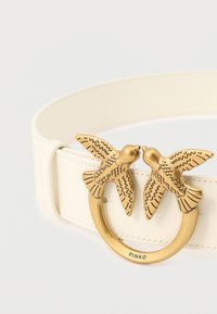 Biały skórzany pasek z okrągłą złotą klamrą przedstawiającą dwie zwrócone do siebie ptaki z grawerowanymi detalami oraz nazwą marki "PINKO" na obręczy.