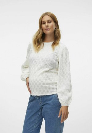 Vit texturerad blus med voluminösa långärmar, ihop med blå jeans. Har en rund halsringning och en figurnära silhuett runt midjan.