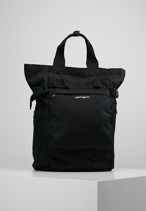 Sac cabas noir Carhartt avec poignées supérieures, poche avant zippée et sangles latérales à boucle sur un piédestal blanc sur fond gris.