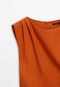 Blouse sans manches orange brûlé avec encolure ronde et détail plissé près de l'épaule gauche, montrant la texture du tissu et l'étiquette à l'intérieur du col.