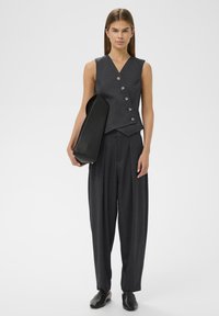 Žena stojí v tmavě šedé bezrukávové vesti s knoflíky a odpovídajících skládacích kalhotách, drží velkou černou psaníčko a má na sobě černé boty.