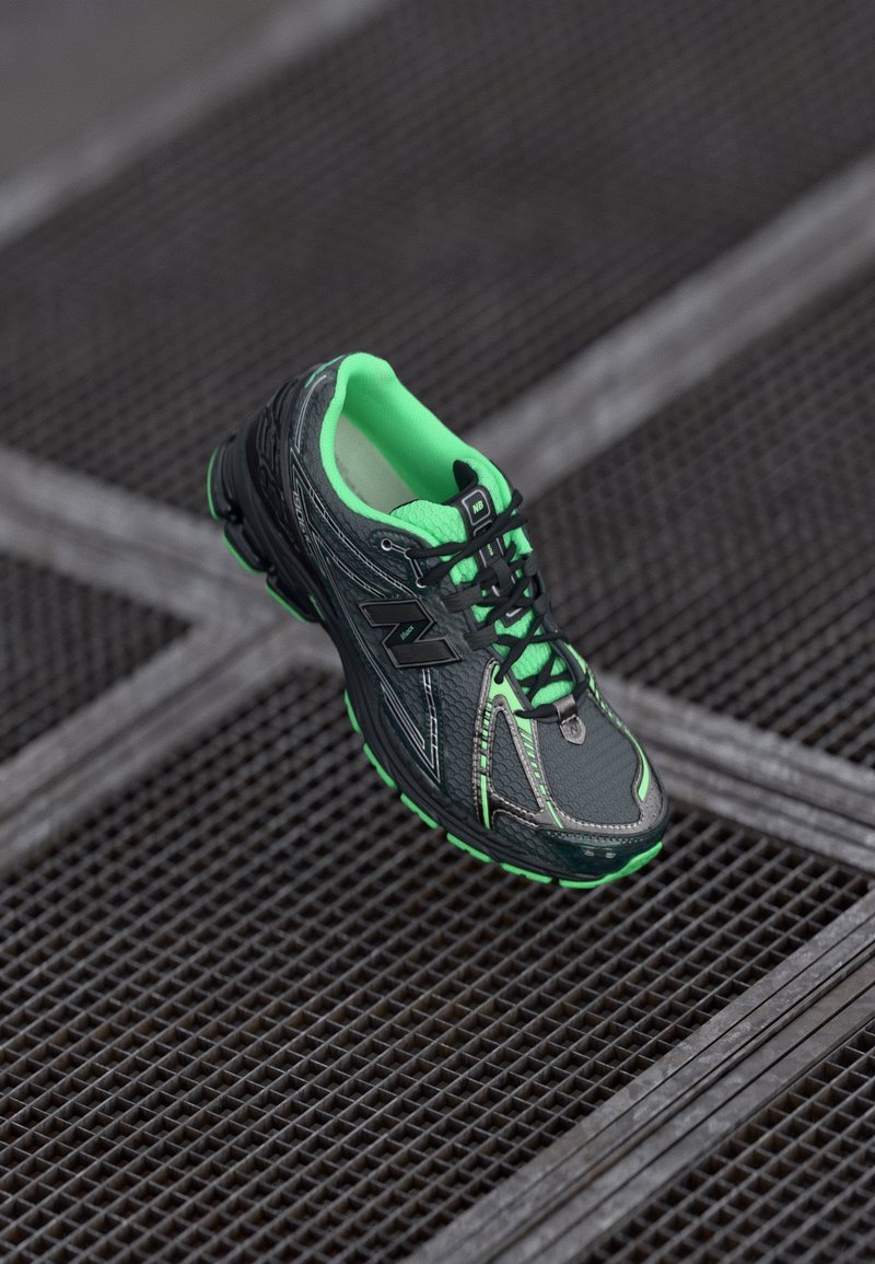 Chaussure de course noire avec des accents verts, superpositions texturées, col rembourré et détails réfléchissants, sur fond de grille en métal.