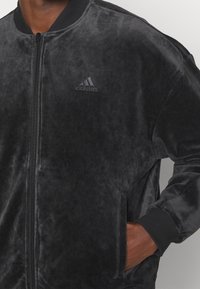 Svart sammet bomberjacka med dragkedja fram, ribbad krage och manschetter. Har subtilt tonat Adidas-logotyp på bröstet och detaljer på fickorna.