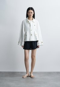 Γυναίκα που στέκεται μπροστά σε απλό τοίχο, φοράει λευκό σακάκι με κουμπιά, μαύρα σορτς και λευκά σανδάλια, κοιτάζει μπροστά με τα χέρια χαλαρά δίπλα στο σώμα.