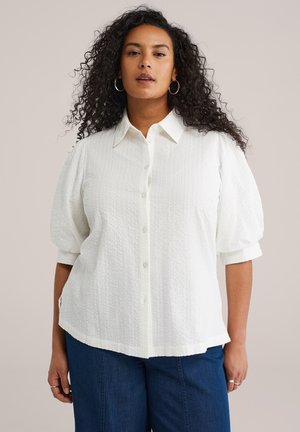 Ženska s kodrastimi lasmi, ki nosi obročne uhane, belo srajco z gumbi in rahlo nabranimi rokavi ter modre kavbojke, stoji pred enostavnim ozadjem.