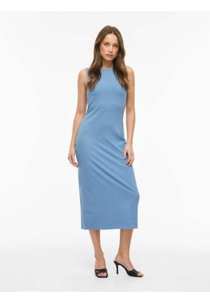 Kobieta stojąca w niebieskiej sukience midi bez rękawów i czarnych otwartych szpilkach na białym tle.