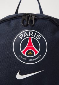 Sac de sport navy présentant un logo rond du Paris Saint-Germain avec un graphique de la Tour Eiffel. Fermeture à glissière avec deux tirettes noires. Swoosh blanc de Nike.
