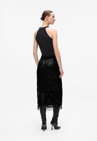 Haut à halter noir associé à une jupe noire en sequins avec des franges en niveaux. Le modèle porte des bottes noires jusqu'aux genoux. Texture lisse et réfléchissante.
