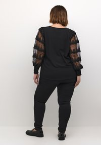 Černý top s průhlednými krajkovými balónovými rukávy, které mají vlnkovaný vzor. Spárováno s černými slim-fit kalhotami a černými plochými botami.