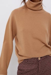 Pull beige à col roulé, coupe oversize, texture douce, col et poignets côtelés. Associé à un pantalon bordeaux à motifs avec des détails de boutons dorés.